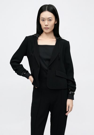 Fekete kapucnis blézer egyedi díszített mandzsettákkal. Strukturált szabás, sima anyag és testhezálló sziluett. Fekete felsővel együtt.