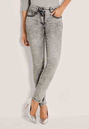 Vrouw draagt grijze acid-wash skinny jeans met een grijze top, staat met één hand in de zak en zilveren hoge hakken.
