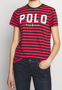 Röd och marinblå randig t-shirt med kort ärm och rund halsringning. Har stor vit text "POLO" och "Ralph Lauren"-logotypen i mitten.