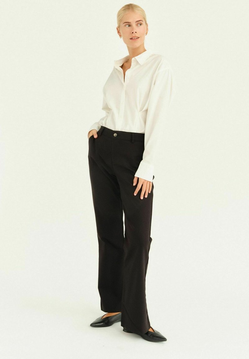 Camicia bianca a maniche lunghe con bottoni abbinata a pantaloni neri a vita alta e scarpe nere basse; presenta un colletto arrotondato e una vestibilità rilassata.
