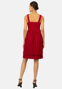 Robe plissée rouge avec un corsage smocké, des bretelles larges et une jupe superposée. La vue de dos montre un haut ajusté avec une section inférieure fluide.