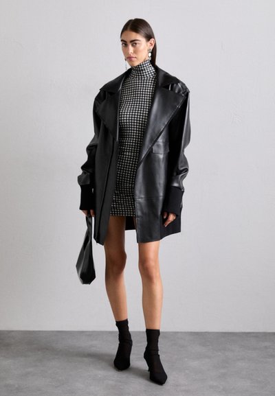 Blazer oversized in pelle nera con rever prominenti, abbinato a un mini abito con motivo houndstooth. Il modello tiene una borsa nera abbinata.