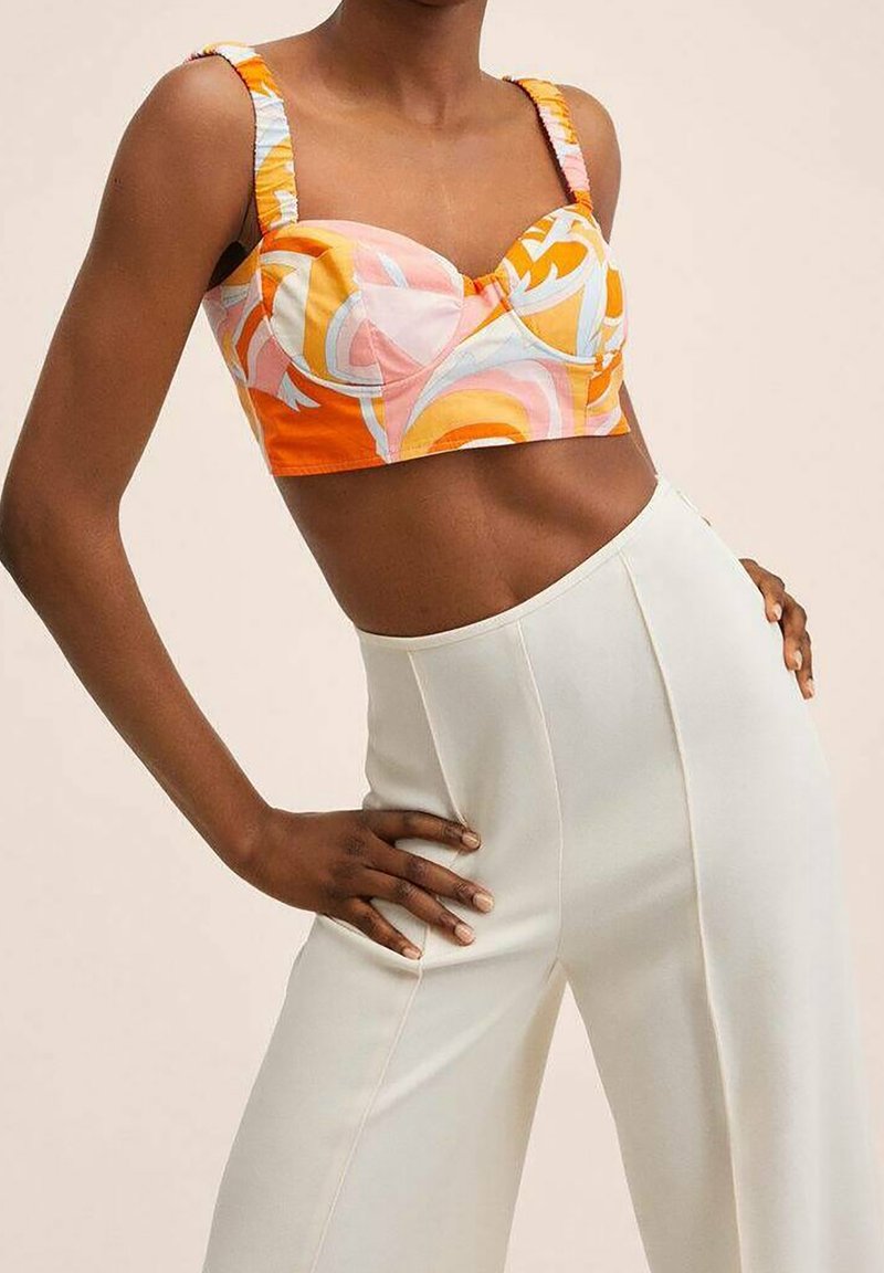 Femme portant un crop top coloré orange et rose à motifs avec un pantalon blanc taille haute, mains posées sur les hanches devant un fond uni.