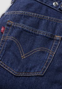 Mørkeblå denimlomme med kontrasterende guldtråd, der har et karakteristisk buet design og et rødt logo-mærke syet ind i stoffet.