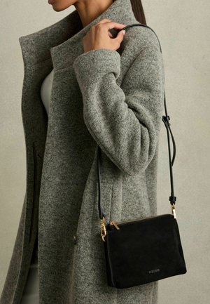 Schwarze Wildleder-Crossbody-Tasche mit einem goldenen Reißverschluss und Metallteilen, die an einem schlanken Tragegurt hängt, neben einem strukturierten grauen Mantel.