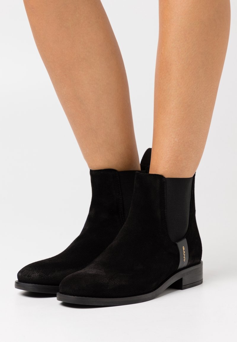 Bottes basses en daim noir avec bout arrondi, panneaux latéraux élastiques, petit talon et accent logo doré sur le côté. Texture lisse dans l'ensemble.