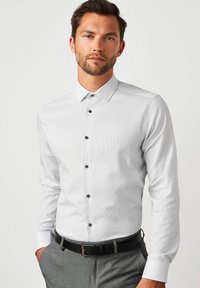 Weiß gestreiftes Hemd mit Knopfleiste, umlegtem Kragen, abgerundetem Saum und schwarzen Knöpfen. Kombiniert mit grauen Hosen und einem schwarzen Gürtel.