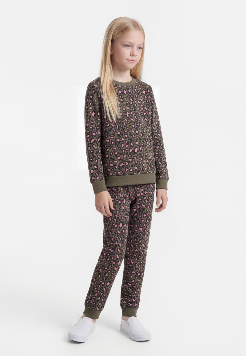 Jong meisje staat, draagt een bijpassend olijfgroen en roze luipaardprint sweatshirt en broek met witte instapschoenen.