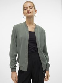 Femme portant une veste bomber zippée gris clair-vert, un haut noir et un pantalon noir, posant contre un fond blanc avec une expression neutre.