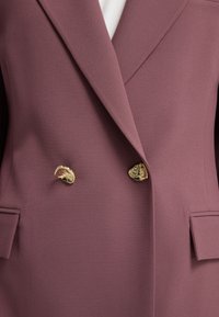 Dubbelzijdige blazer in een stoffige rozenkleur, gemaakt van een zachte stof. Beschikt over gouden knopen en twee zijzakken. Strak ontwerp met scherpe revers.