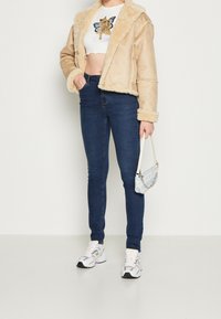 Béžová bunda z umělé ovčí vlny se širokým límcem, doplněná o bílý krátký top, tmavě modré úzké džíny a šedé sportovní tenisky.