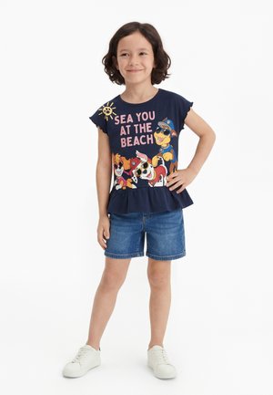 Barn med kort krøllet hår har på en mørkeblå skjorte med "Sea you at the beach" og tegneseriehunde, denimshorts og hvide sneakers, smilende.