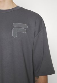Camiseta de manga corta gris con cuello redondo, que presenta un diseño gráfico texturizado en la parte izquierda del pecho. Acabado de tela suave.