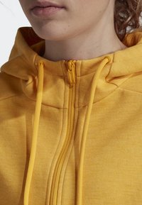 Žlutá mikina na zip s texturovanou látkou, vybavená odstínovým zipem a stahovacími šňůrkami, a s přiléhavou kapucí pro těsné přizpůsobení.