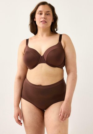 Kvinde med medium hudtone iført brun bh med fuld dækning og matchende højtaljede trusser, stående mod en ensfarvet lys baggrund.