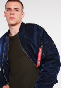 Sötétkék bomber dzseki fényes anyagból, narancssárga cipzárral, bordázott mandzsettával és egy mellzsebből kiemelkedő piros "REMOVE BEFORE FLIGHT" felirattal.