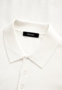 Polo blanc côtelé avec un col classique et trois boutons, comportant une étiquette noire "Matinique" à l'intérieur de l'encolure.