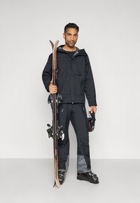 Giacca da sci impermeabile nera con tasca con zip, abbinata a pantaloni da sci neri con pannelli grigi. Il modello tiene in mano sci in legno e un casco.