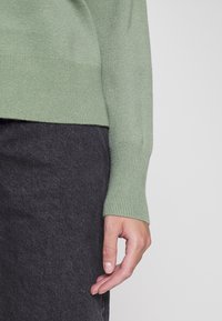 Hellgrüner Strickpullover mit gerippten Bündchen, kombiniert mit einem dunkelgrauen Rock. Strukturierter Stoff mit weichem Finish, zeigt eine locker resting Hand.