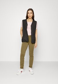 Colete preto acolchoado sem mangas sobre uma camisa justa de cor rosa claro, combinado com calças verde-oliva e sapatilhas brancas com detalhes em vermelho.
