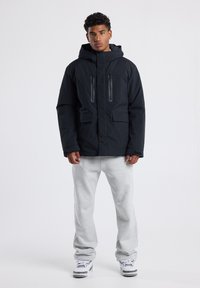 Chaqueta negra acolchada con capucha, dos bolsillos de pecho con cremallera y un ajuste en la parte inferior. Llevada con pantalones de chándal grises y zapatillas blancas.