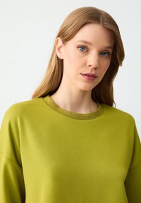Sweatshirt vert avec un col métallisé texturé doré, offrant une coupe décontractée et un tissu doux. Le design est simple et minimaliste.
