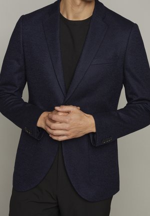 Uomo che indossa un blazer blu scuro sopra una camicia nera, mani giunte all’altezza della vita, su uno sfondo neutro uniforme.