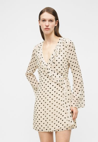 Γυναίκα που φοράει φόρεμα τύπου wrap σε κρεμ απόχρωση με μαύρα μοτίβα καρδιών και βολάν στα τελειώματα, στέκεται μπροστά σε απλό λευκό φόντο.