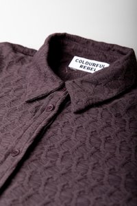 Camicia texturizzata di un oscuro colore viola con colletto, caratterizzata da un motivo geometrico, due bottoni di chiusura e un'etichetta con la scritta "COLOURFUL REBEL."
