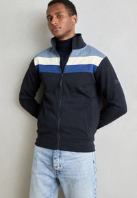 Jachetă navy cu fermoar, cu dungi orizontale de culoare albastru deschis, crem și albastru închis pe piept; material din amestec de bumbac; două buzunare frontale.