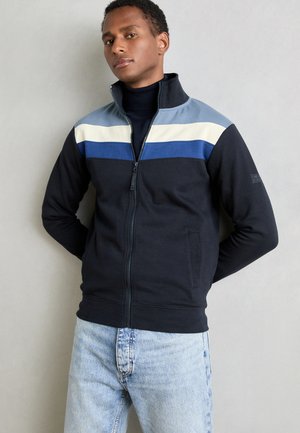 Chaqueta de chándal azul marino con franjas horizontales en celeste, crema y azul oscuro en el pecho; material de mezcla de algodón; dos bolsillos frontales.