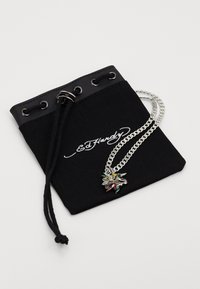 Colar de corrente prateada com um pendente multicolorido descansando sobre um saco preto com cordão, apresentando um logótipo branco "Ed Hardy."