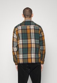 Muž v zeleno-oranžovo-bílo-černé kostkované bundě a černých kalhotách, stojící zády k bílému jednobarevnému pozadí.