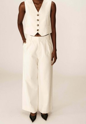 Débardeur blanc court avec quatre grands boutons marron et un pantalon large taille haute plissé, associé à des escarpins noirs pointus.