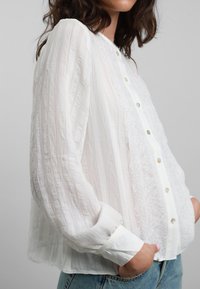 Camicia bianca in tessuto testurizzato, con un pannello in pizzo e chiusura a bottoni. Vestibilità ampia, maniche con polsini e una leggera lucentezza. Design leggero e arioso.