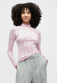 Jauna sieviete ar cirtainiem matiem, tērpusies rozā ar garām piedurknēm ziedu raksta topā un gaiši pelēkos ziedu raksta biksēs, rokas kabatās, skatās taisni priekšā.