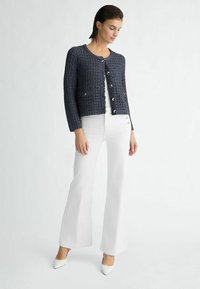 Marineblauwe tweed cardigan met een ronde hals, lange mouwen, knoopsluiting en twee voorzakken, gecombineerd met witte flared broek en witte hakken.