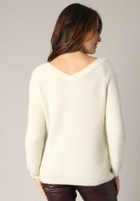 Pull en tricot blanc avec un tissu texturé, décolleté en V dans le dos, et manches longues, associé à un pantalon marron foncé.