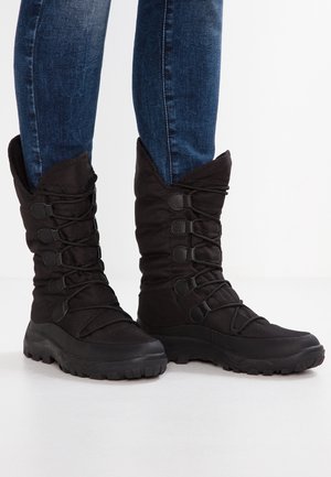 Bottes d'hiver noires mi-mollet avec semelles épaisses et plusieurs œillets, portées avec un jean skinny bleu foncé sur fond blanc.