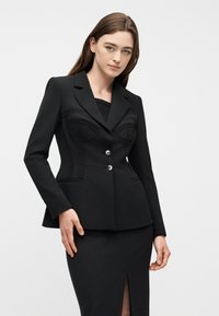 Νεαρή γυναίκα με ίσια καστανά μαλλιά φοράει εφαρμοστή μαύρη σακάκι με δομημένες ραφές και ταιριαστή μαύρη φούστα με σκίσιμο μπροστά.
