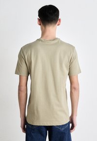 Beige bavlněné tričko s krátkým rukávem, kulatým výstřihem a hladkou texturou, nošené s modrými džíny, pohled zezadu.