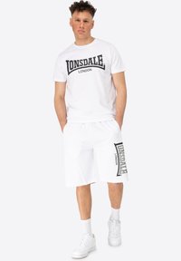 Bijela pamučna majica s crnim natpisom "LONSDALE LONDON", uparenim s bijelim kratkim hlačama koje imaju bočni logo. Bijele tenisice upotpunjuju izgled.