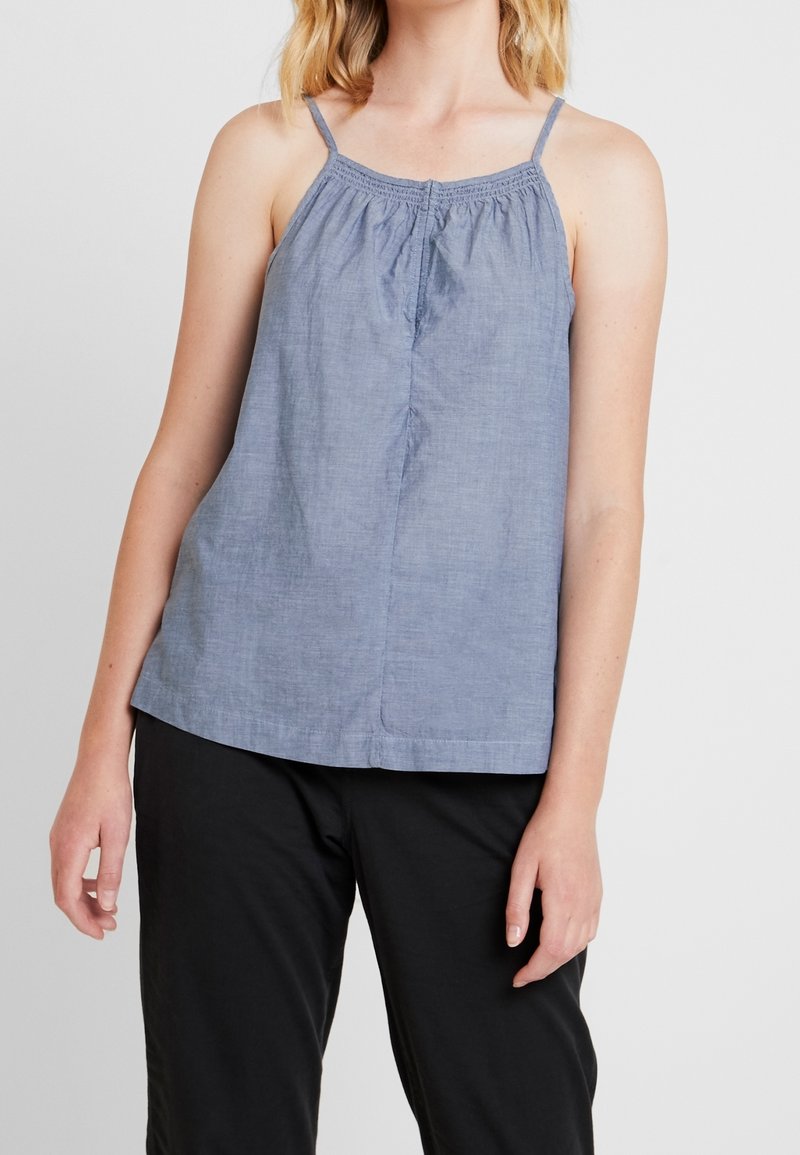 Femme portant un haut en chambray bleu sans manches avec des bretelles fines et un pantalon noir sur un fond uni.