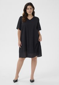 Black, knee-length dress with short sleeves, featuring a perforated design. V-neckline and loose fit for comfort. Worn with black mules.