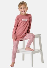 Roze tuniek met grafische print "CAT", gecombineerd met rood-wit gestreepte leggings. Kind poseert op een witte kruk met blote voeten.