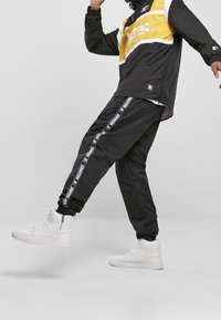 Schwarze und gelbe Windjacke mit weißen Streifen, kombiniert mit schwarzen Jogginghosen mit seitlichem Branding und Beinbündchen, getragen mit weißen High-Top-Sneakern.