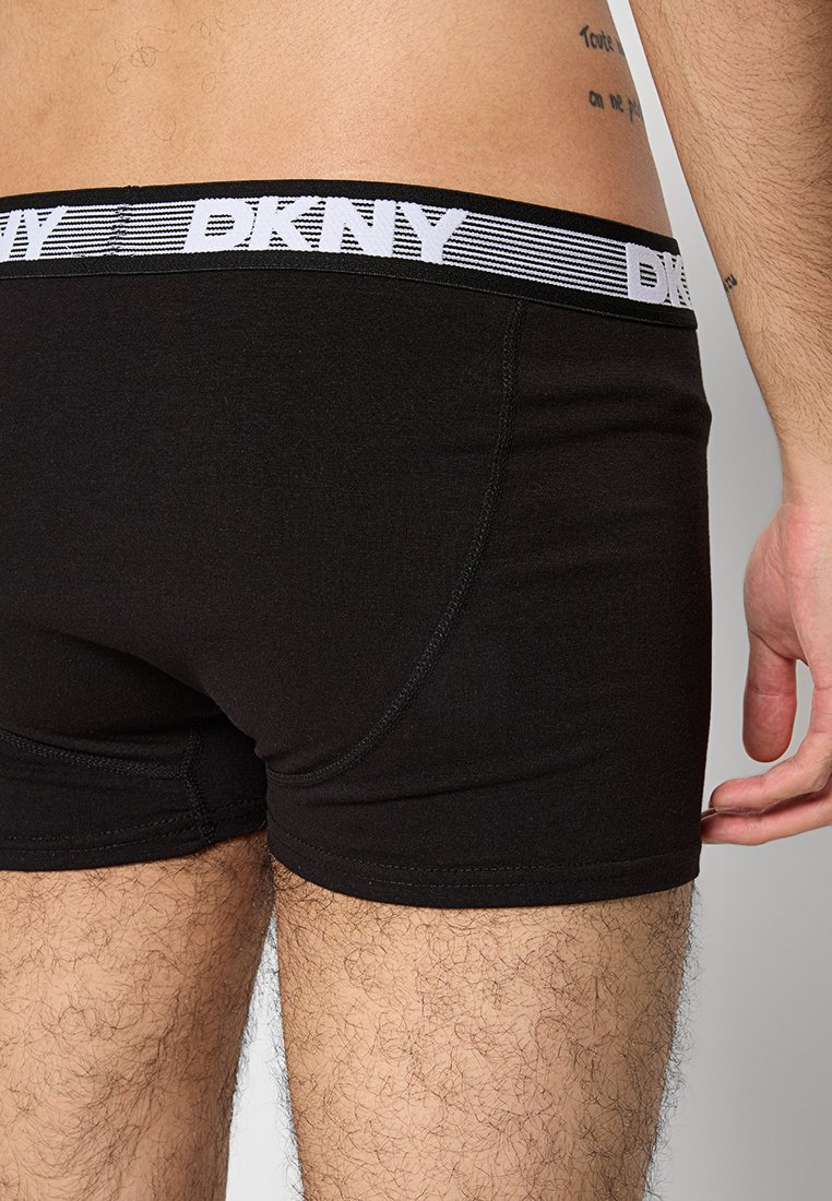 Męski tors i nogi w czarnych bokserkach DKNY z markowym pasem, ukazujące boczną i tylną stronę z częściowym tatuażem na dolnej części pleców.