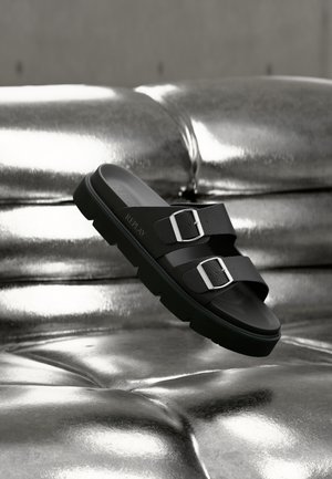 Black slide sandals with dual buckles, textured straps, a thick sole, and a smooth finish, resting above a metallic surface.