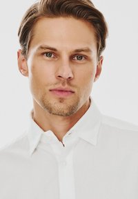 Biała koszula z kołnierzykiem, wykonana z gładkiego materiału. Model ma krótkie, jasnobrązowe włosy i jasne zarosty. Neutralne tło.