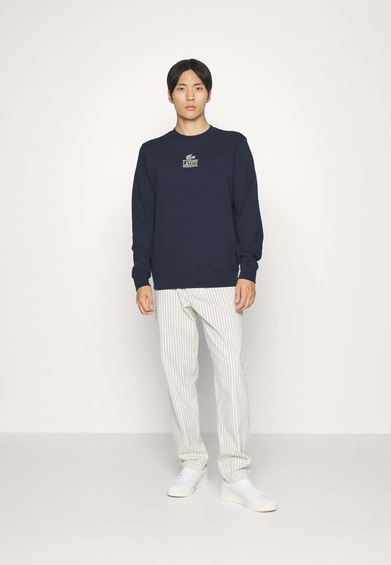 Granatowa bluza z okrągłym dekoltem i małym logo Lacoste, w zestawie z białymi spodniami w zielone paski i białymi trampkami. Prosty design.